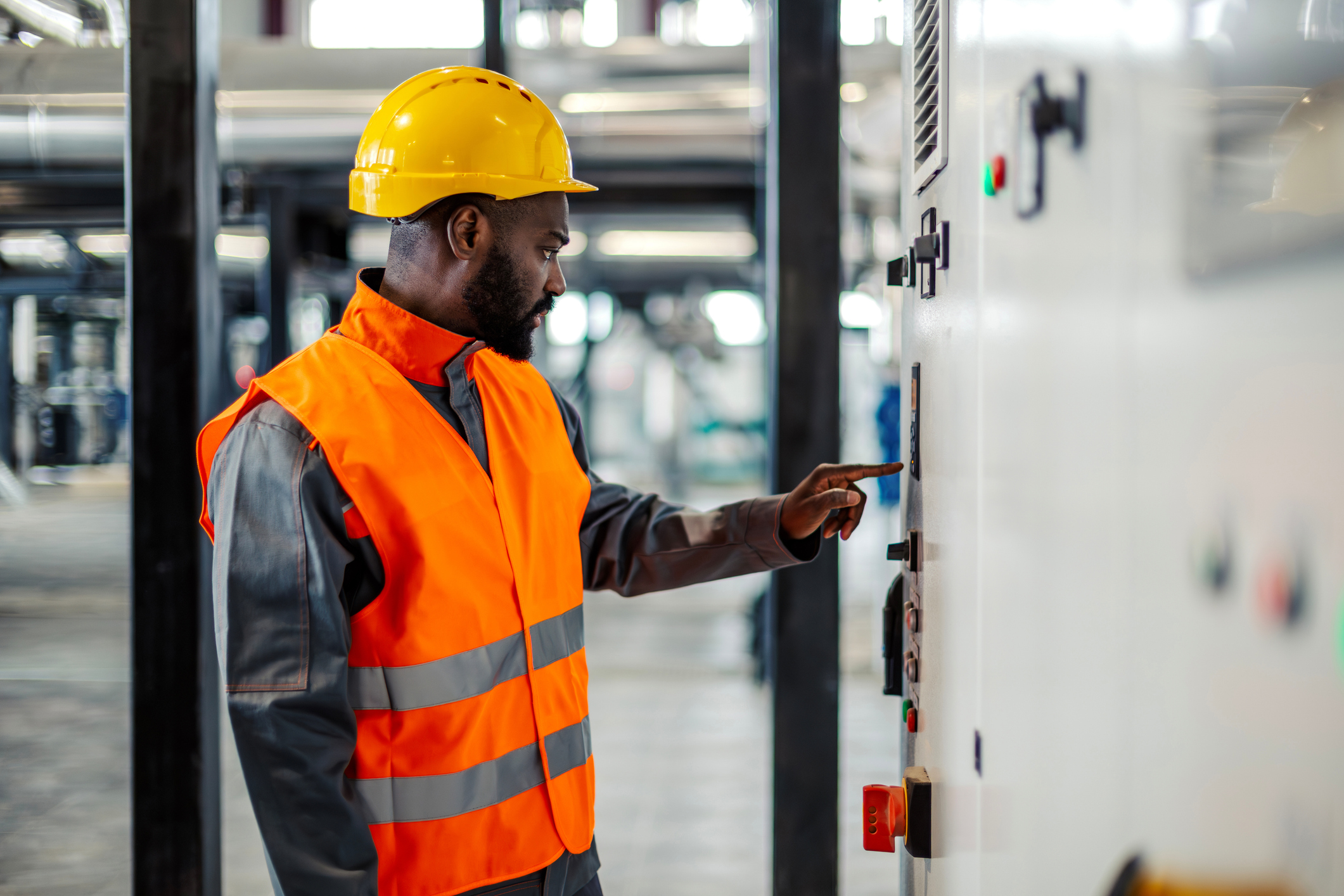 Focused male engineer wearing a hard hat and high-visibility vest is operating a control panel, ensuring safety and efficiency in an industrial factory setting